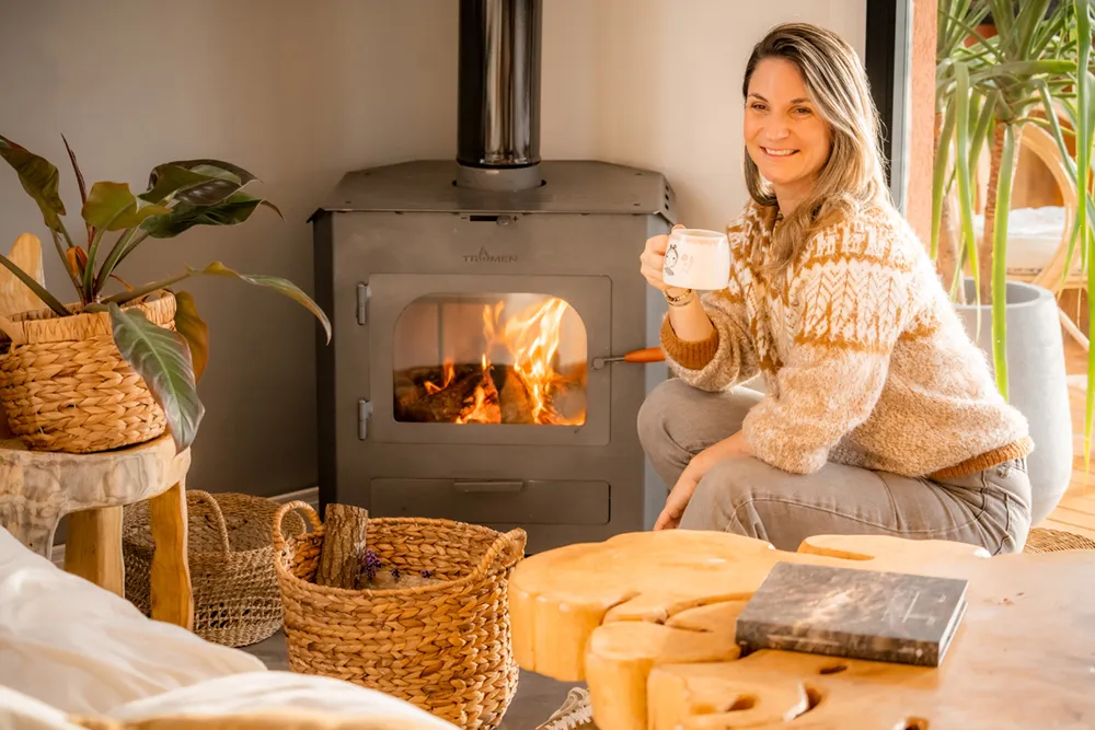 A woman sits drinking coffee by the fireplace.